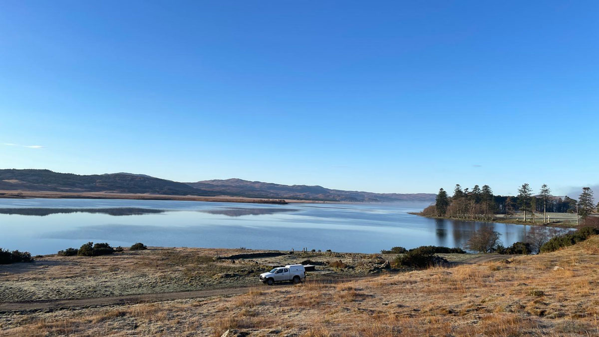 A lake, surrounded by low hills and trees, is overlooked from a nearby hill. In the mid-ground, a white truck drives across the frame.