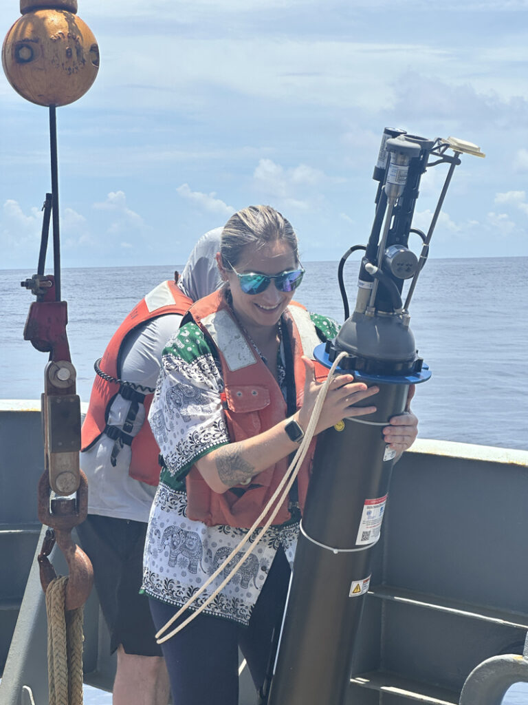 A researcher stands on the outside deck of a large ship, holding a large gray cylindrical scientific instrument that is slightly taller than them. The researcher wears sunglasses, a T-shirt and an orange safety vest. The metallic cylinder is outfitted with knobs, tubes, and a variety of gauges.