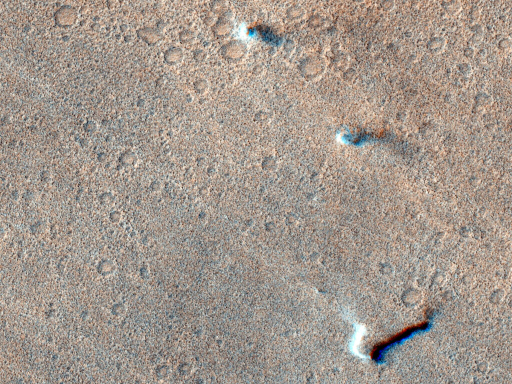 Three dust devils on the Martian surface, seen from above and appearing as small white splotches.