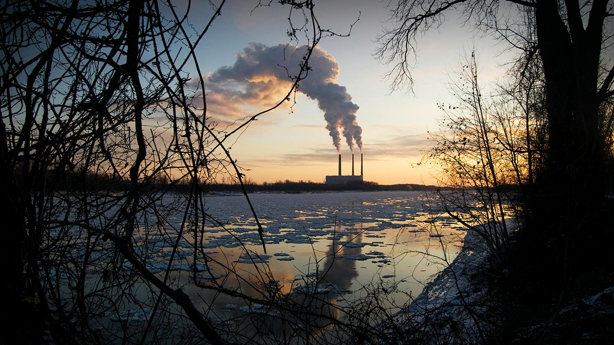 Emissions billow from a power plant’s smokestacks in the distance beyond a partially ice-covered river.