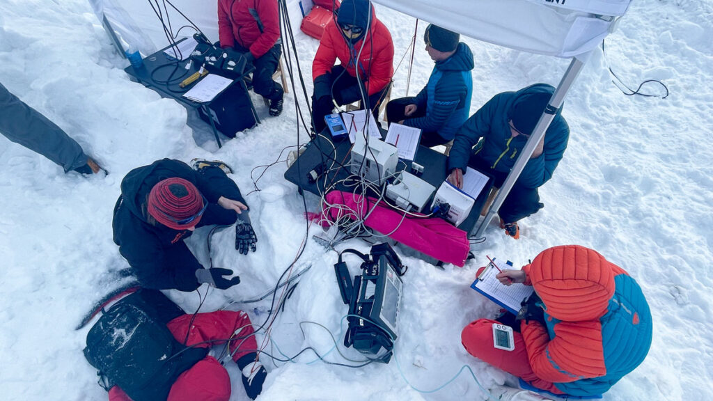 An overhead image of six people in winter jackets under a canvas tent. Medical devices and cables snake through the snow. A participant in a red jacket lies face down, half buried in snow.