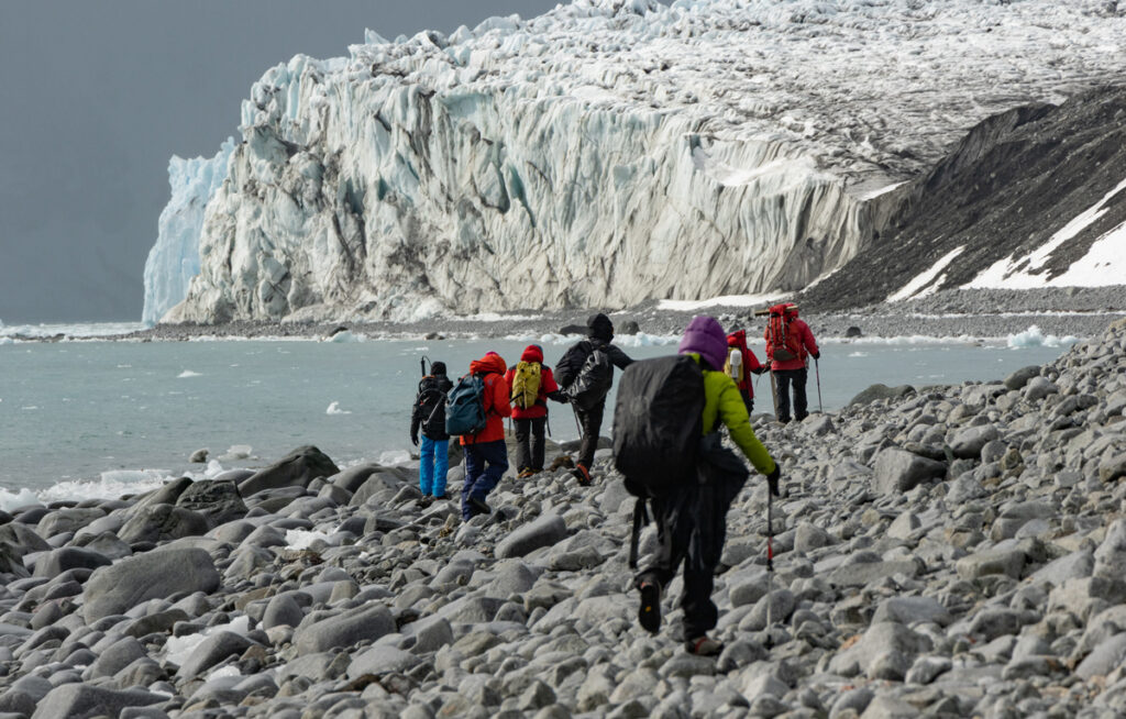 A group of researchers walks across a rocky Antarctic beach toward a towering glacier.