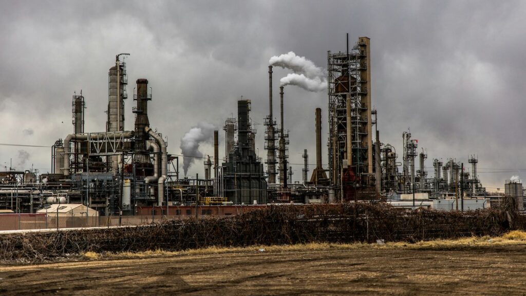 Smokestacks at an industrial facility release gases into the air under a cloudy sky.