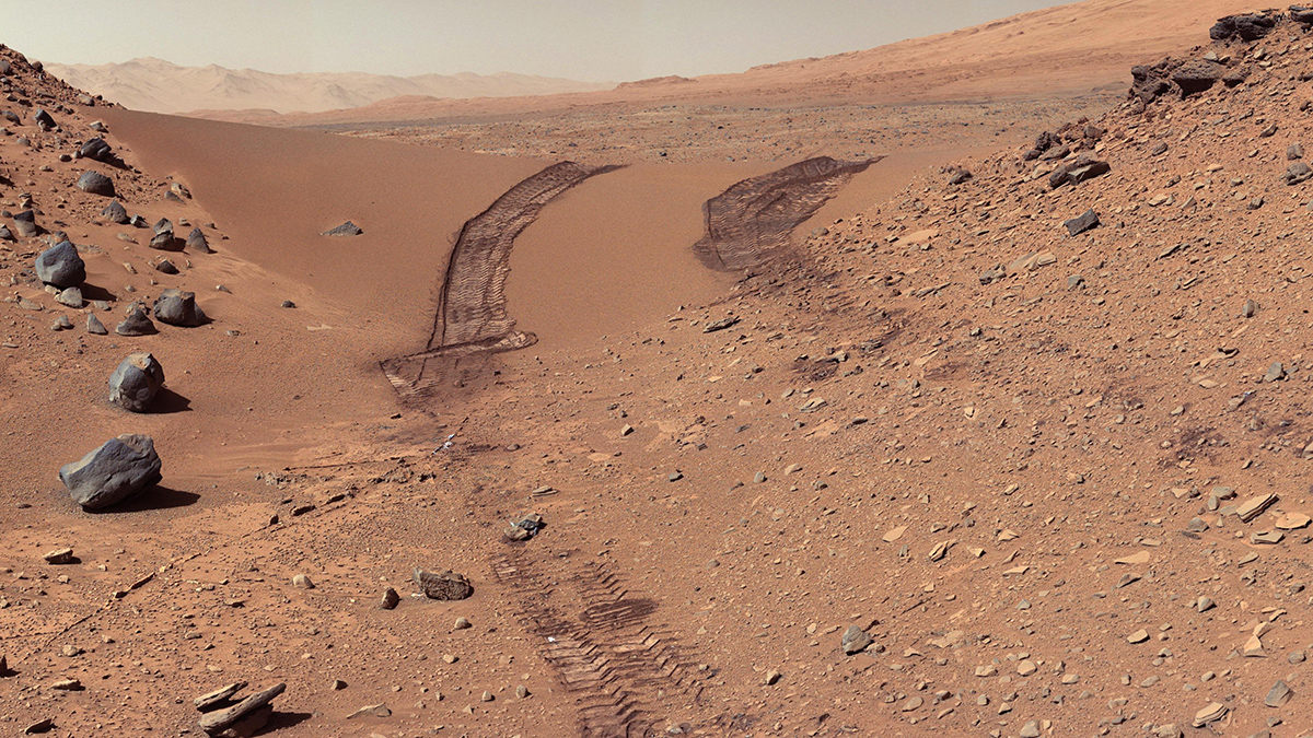 Image taken by Mars Curiosity Rover of the Martian surface showing its parallel wheel tracks (center, spaced 9 feet apart) in red Martian regolith. In the center of the image, the regolith appears to be fine grained. On both sides, slopes are studded with boulders and cobbles of varying shades of red and gray black. The regolith in the foreground is scattered with many angular red or gray cobbles. The gray Martian sky forms the backdrop with red hills in the distance.