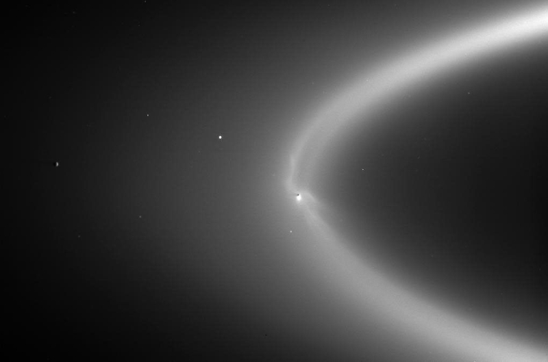 A grayscale image of a section of Saturn’s E ring, which is backlit and appears diffuse and wispy. Enceladus and its south pole plumes are embedded within the ring.