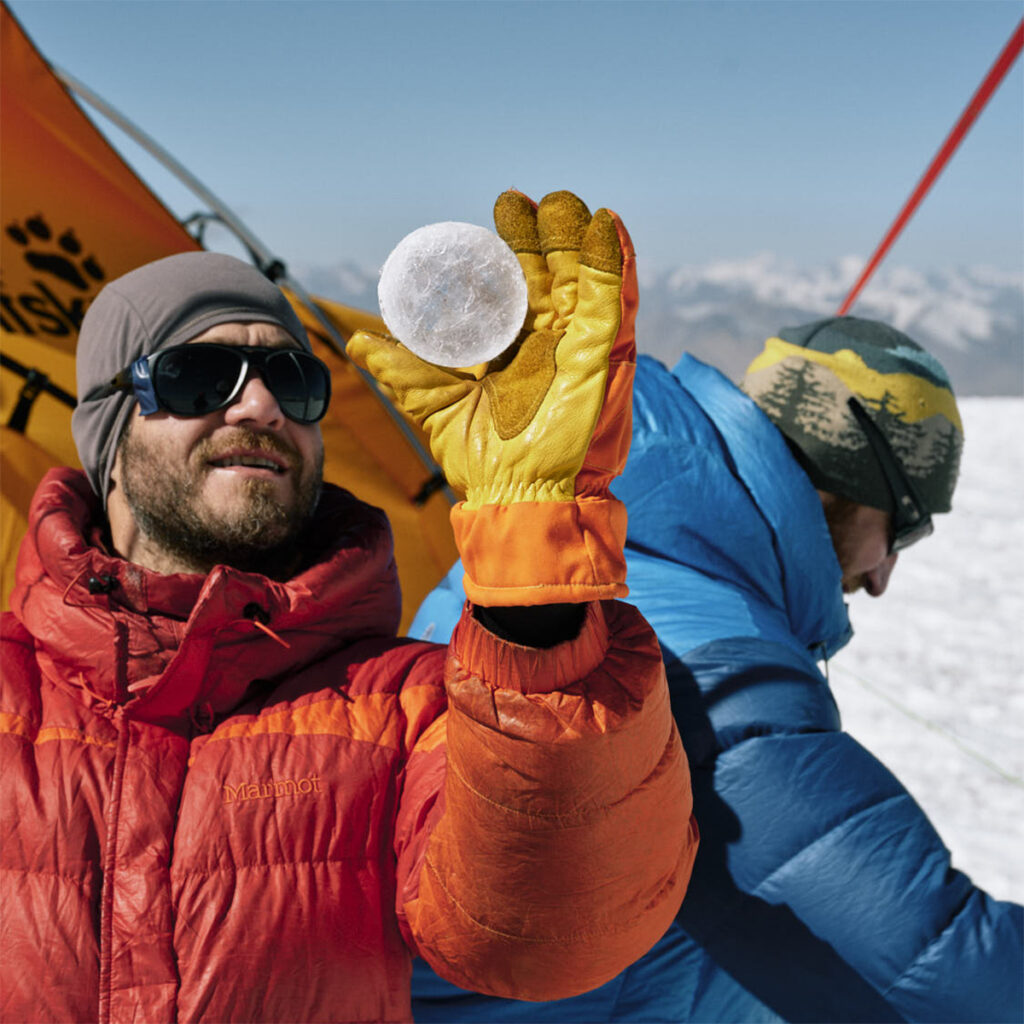 A researcher wearing an orange coat and sunglasses holds an ice cylinder up as if to look through it.