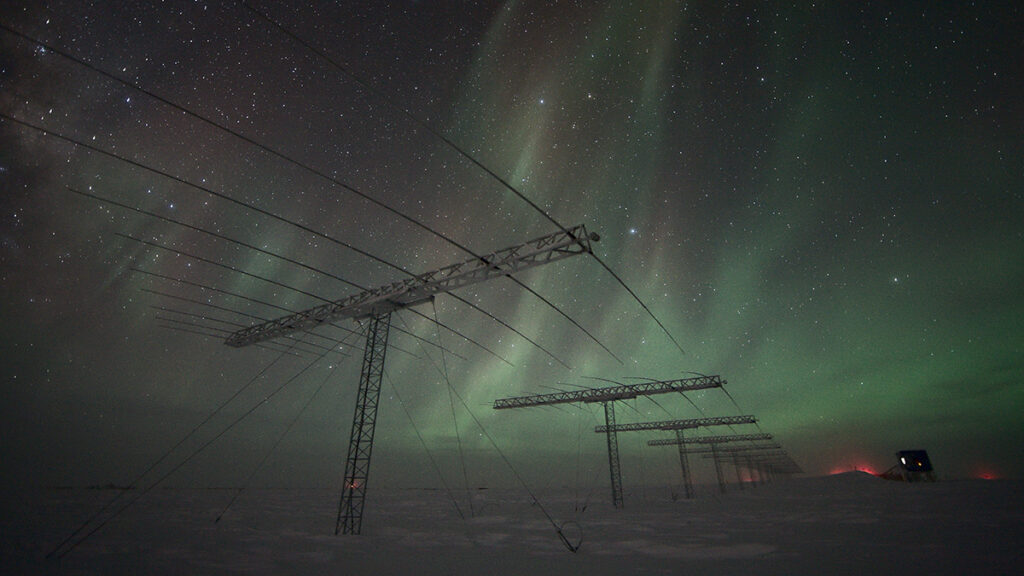 A series of structures that look like electrical poles extend into the distance on an icy surface. The sky above is full of stars and streaked with green aurorae.