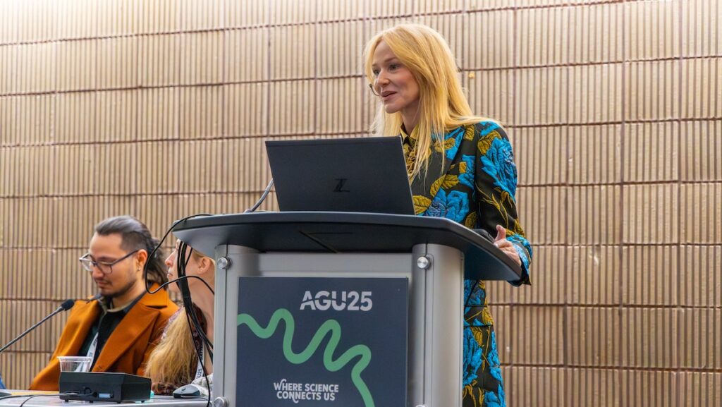 Jewel, a red-headed woman dressed in a blue jacket, speaks at a podium. Two other people are sitting at the table to her right.