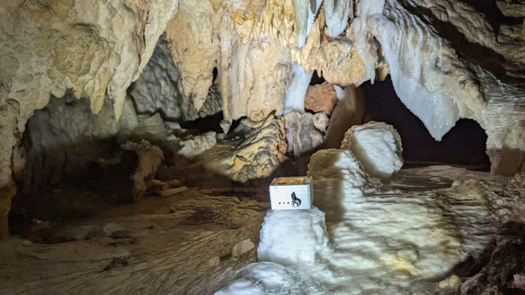 An automated hydrological drip logger (small rectangular box) sits atop a white stalagmite below stalagmites dripping with water in a tight cave space illuminated with bright light.
