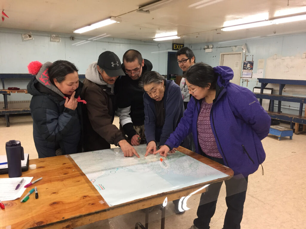 Six people gather around a map, pointing at a spot near the middle.