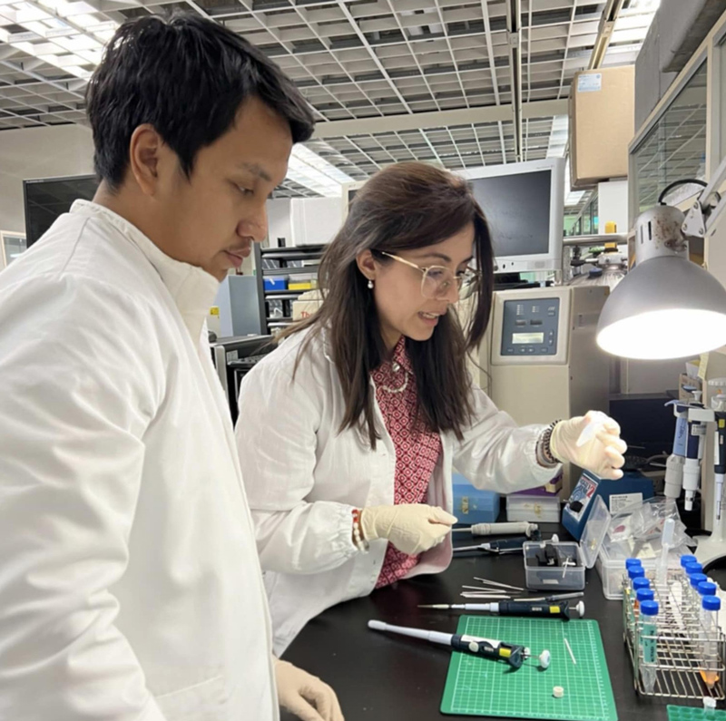 A woman wearing glasses, a white lab coat, and white gloves holds a vial at a workstation while a young man in a white lab coat watches.