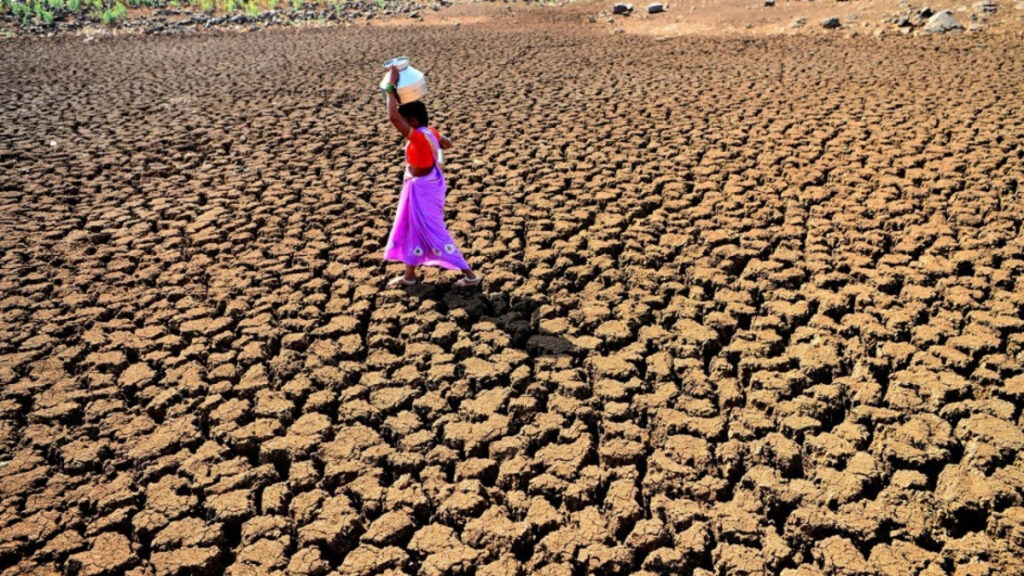 A woman in a red top and purple skirt walks across parched ground carrying a jug of water on her head.