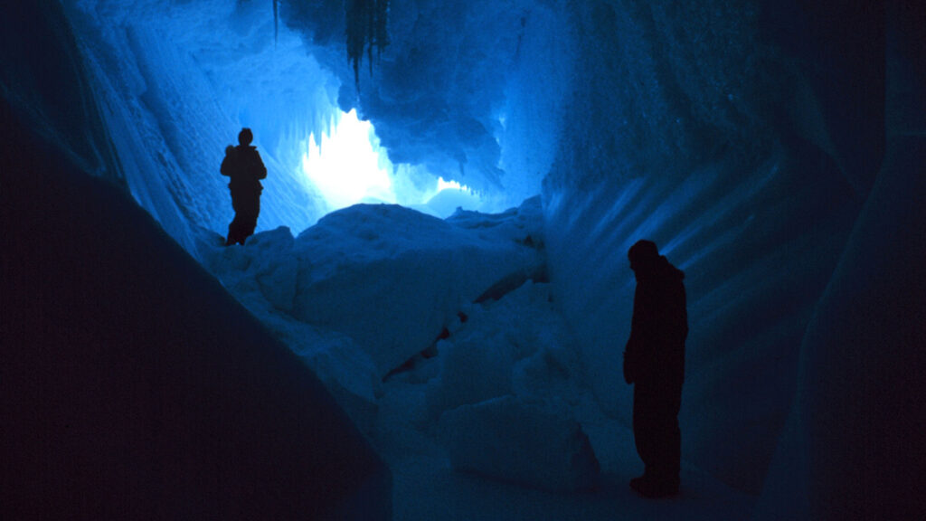 Two people in shadow explore a dark ice cave with ice stalactites and columnar-appearing sides. Light from the entrance backlights the scene.