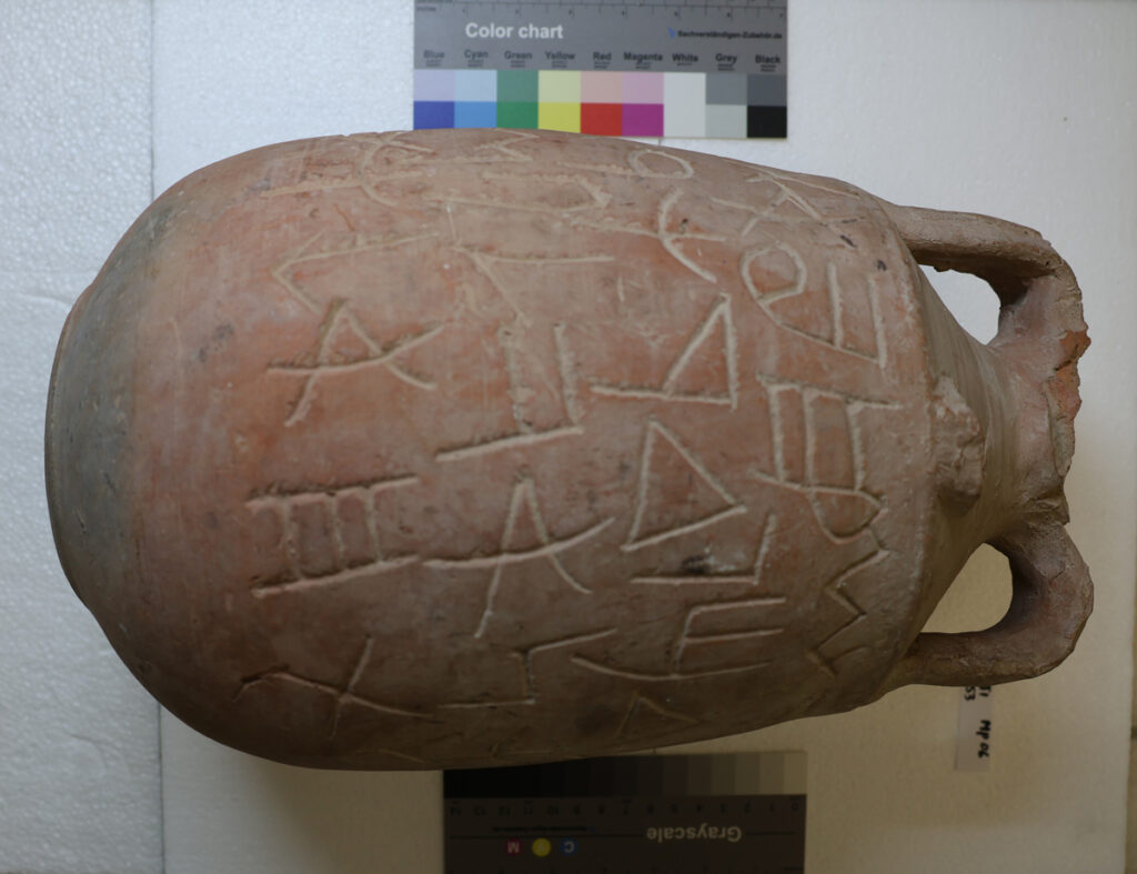 A seemingly ancient terra-cotta-colored pot with a rounded bottom and double handles sits on its side. Phoenician symbols that translate to nonsense are carved into the visible side of the pot; above the pot is a color chart and below it is a grayscale chart.