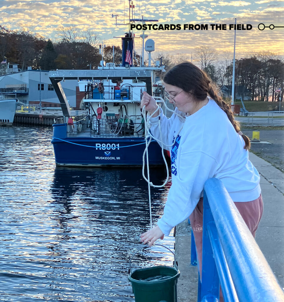 A woman in profile leans over a blue railing on a concrete pier. With her left hand, she reaches toward a green plastic bucket of water that she is holding up by an attached thin white rope held in her right hand and loosely wrapped around her arm. Behind her can be seen the back of a boat with the NOAA logo, “R8001,” and “Muskegon, MI” printed on its stern.