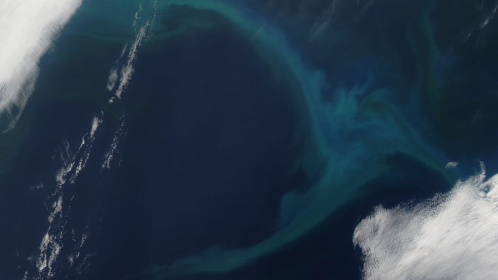 A backward C-shaped swirl of milky blues and greens appears against a deep blue background. In the foreground, toward the edges of the image, clouds appear as white patches.
