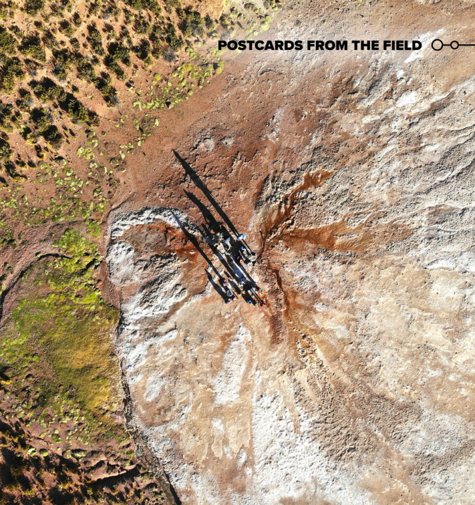 A group of people with instruments are seen from far above, their long shadows extending toward the top left of the image. Around them are various types of terrain, from a dry looking expanse of tan soil with white areas to richer looking reddish soil with green objects that appear to be plants.