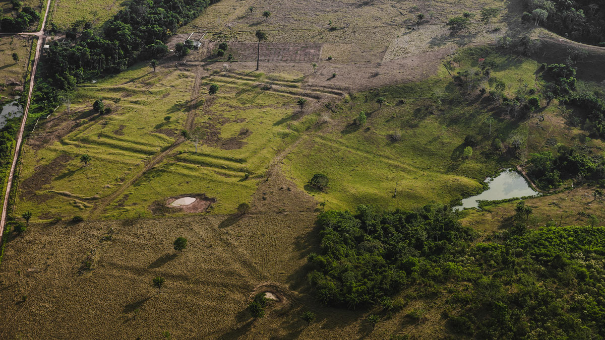 An aerial image shows a green, grassy area where there are large rectangular indentations in the ground. Trees are visible on either side of the frame, and a road is visible on the left.