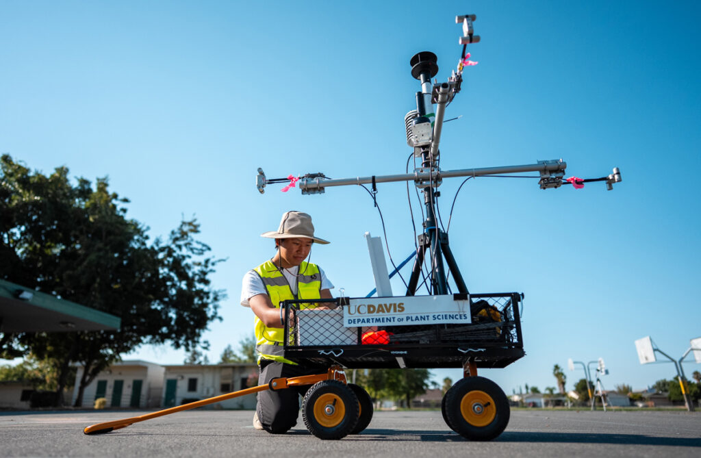 A researcher in safety vest and sun hat pulls a wheeled cart equipped with a tall pole supporting multiple weather sensors and monitoring equipment, branded with UC Davis Department of Plant Sciences.