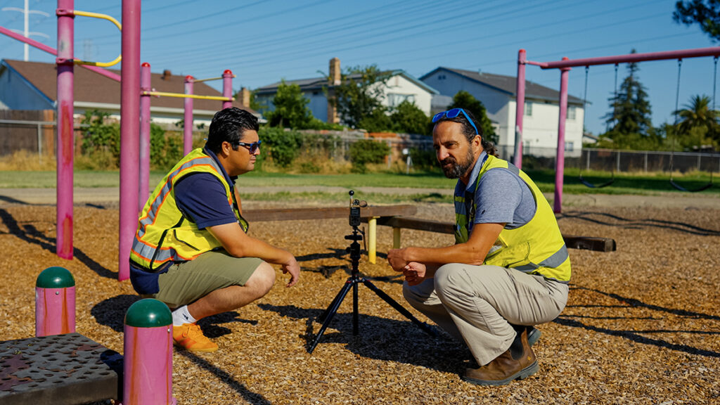 Two researchers in safety vests crouch on a wood chip playground surface, examining a tripod-mounted environmental sensor positioned between playground equipment.