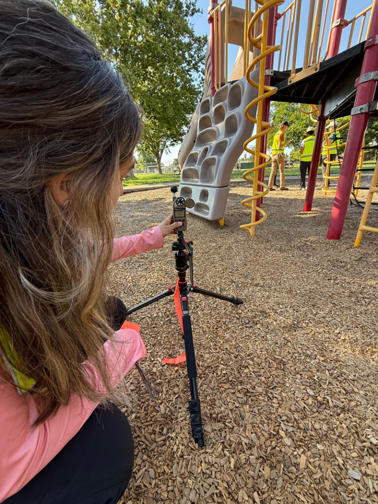 A researcher with long hair wearing a pink jacket holds a handheld environmental sensor on a tripod while standing on wood chips near colorful playground equipment.