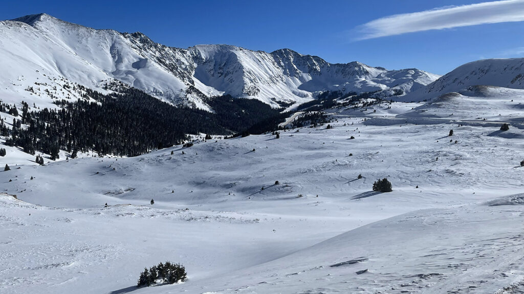 A windswept, snow-covered alpine pass with mountains in the background under a blue sky