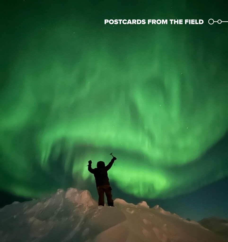 A person silhouetted against a green swirling aurora stands atop a pile of snow and ice, holding up both hands. In the person’s right hand is a rock hammer.