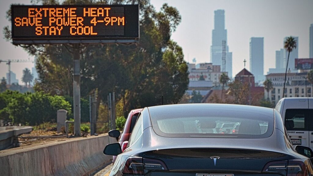 A sign over a busy highway reads “Extreme heat, save power 4–9 p.m., stay cool.”