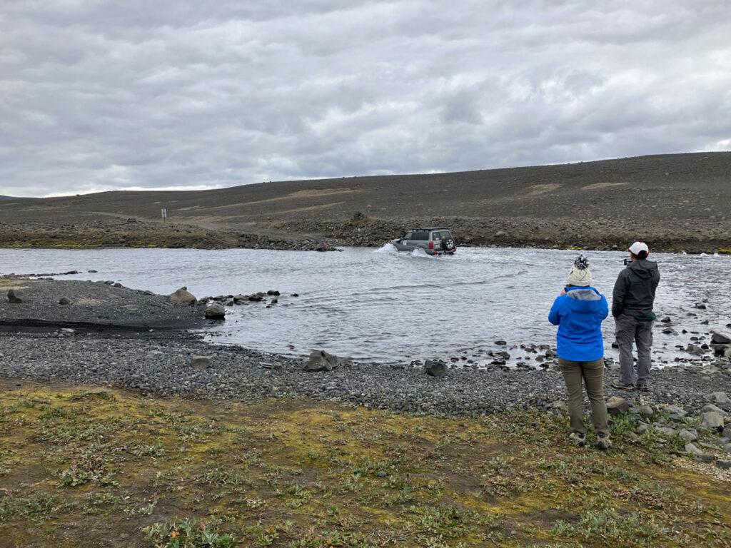 An offroad vehicle drives across a wide but shallow stream in a rocky landscape as two people look on from the near shore.