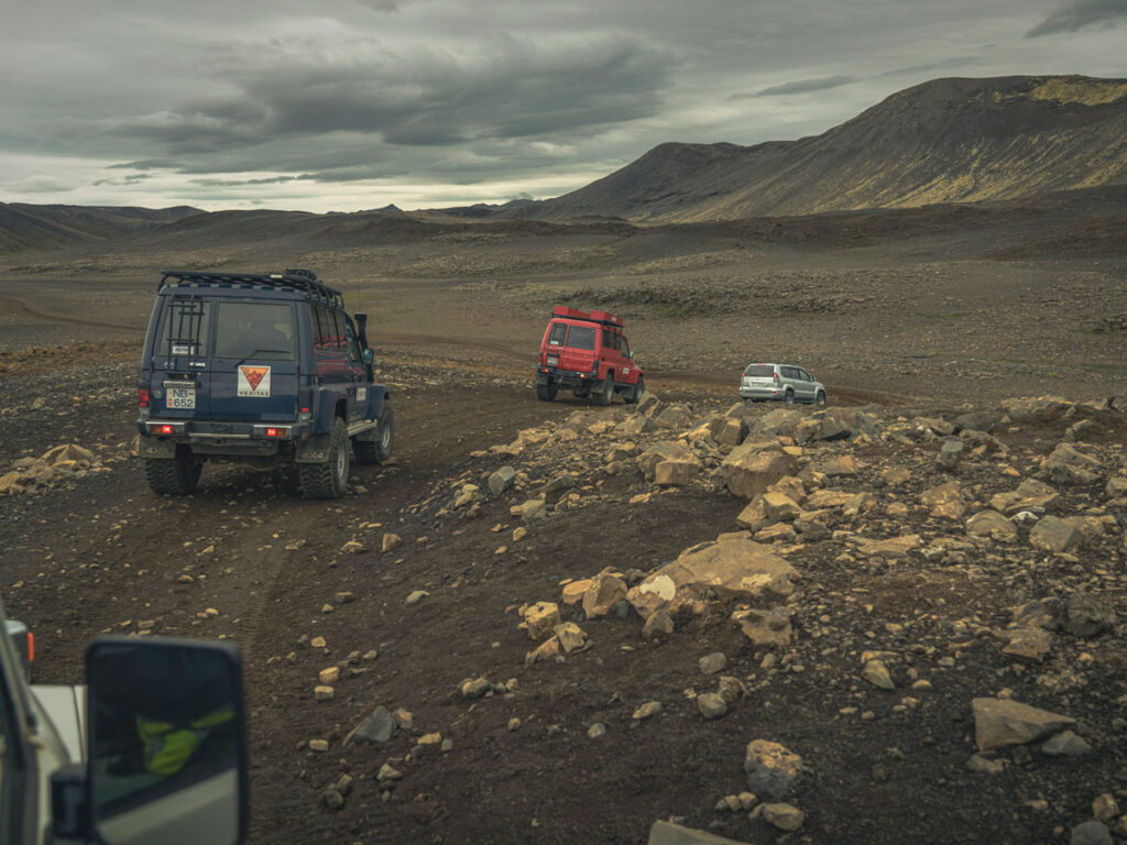 View from out the front passenger window of a vehicle showing three offroad trucks driving single-file amid a wide expanse of dark rock in a barren landscape and under dark clouds.
