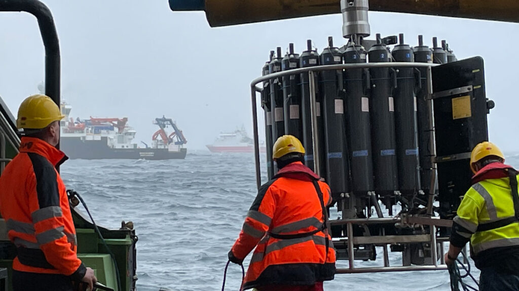Three people in bright yellow and orange jackets appear to be on a boat. They are reaching for a collection of tanks that will be lowered into the water. Two other ships are visible in the distance.