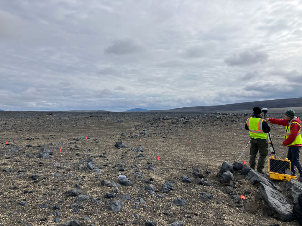 Two researchers wearing yellow safety vests stand near a tripod mounted with scientific equipment amid a wide open expanse.
