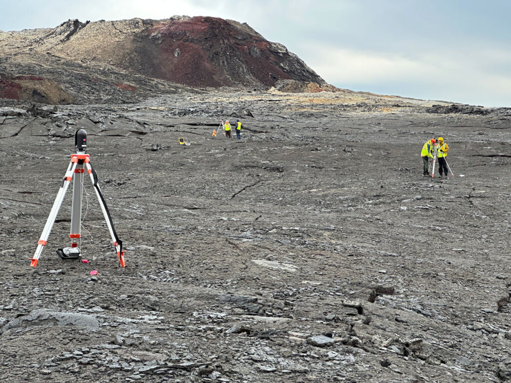 Several tripods holding scientific equipment and spaced far apart stand on gray rocky ground. Two pairs of scientists stand near two of the tripods in the distance.