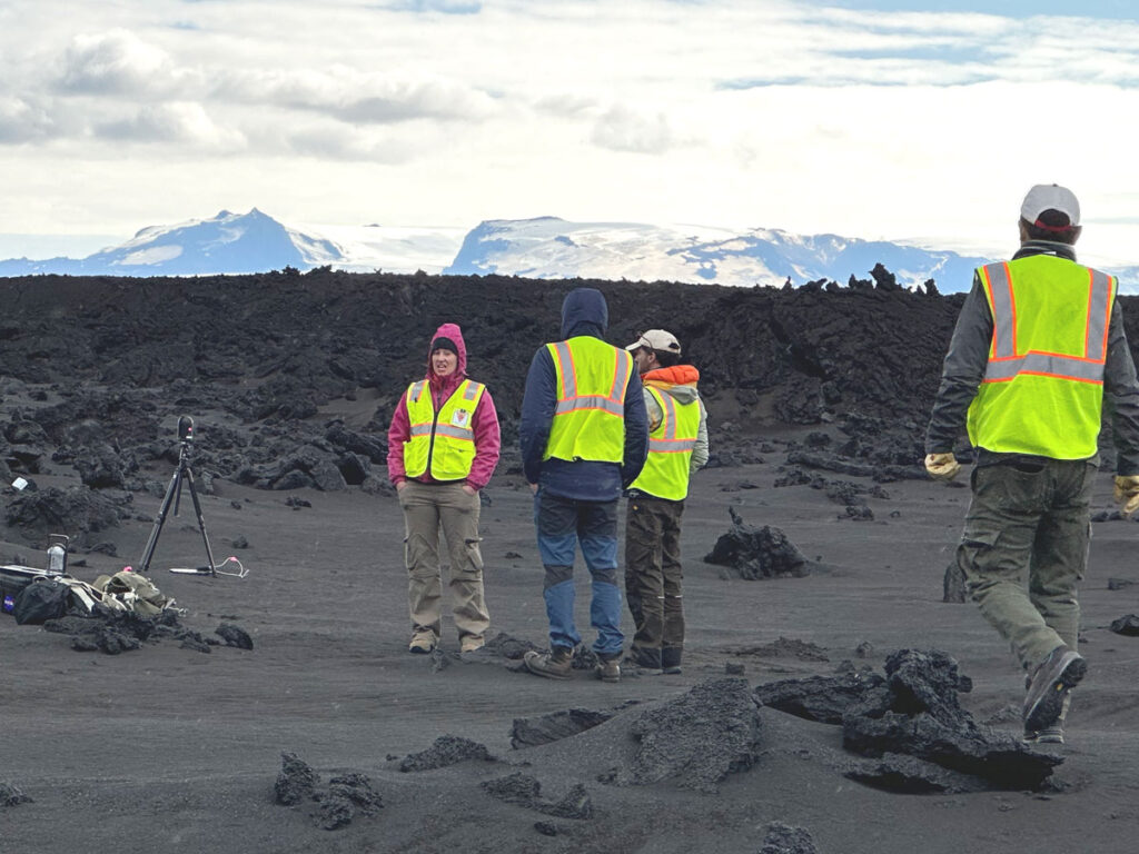 Several scientists wearing yellow safety vests stand and talk amid a dark rocky landscape, with scientific equipment set up nearby.