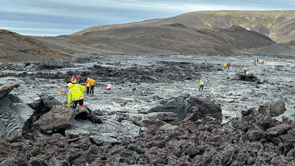 Scientists wearing bright yellow safety vests stand in various places amid an expanse of dark volcanic rock with barren hills in the background.