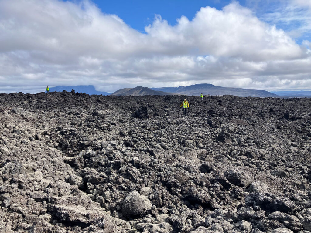 Three scientists wearing yellow safety vests walk in the distance across an area covered by jagged, blocky dark rocks.