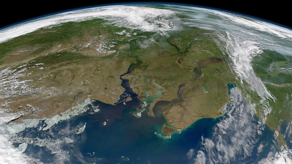 A satellite view of a portion of Earth shows the planet’s curved horizon at the top of the image. Green and brown landmasses are cut by two large, blue, winding rivers that empty into the ocean in the foreground. Clouds are visible on the edges of the image.