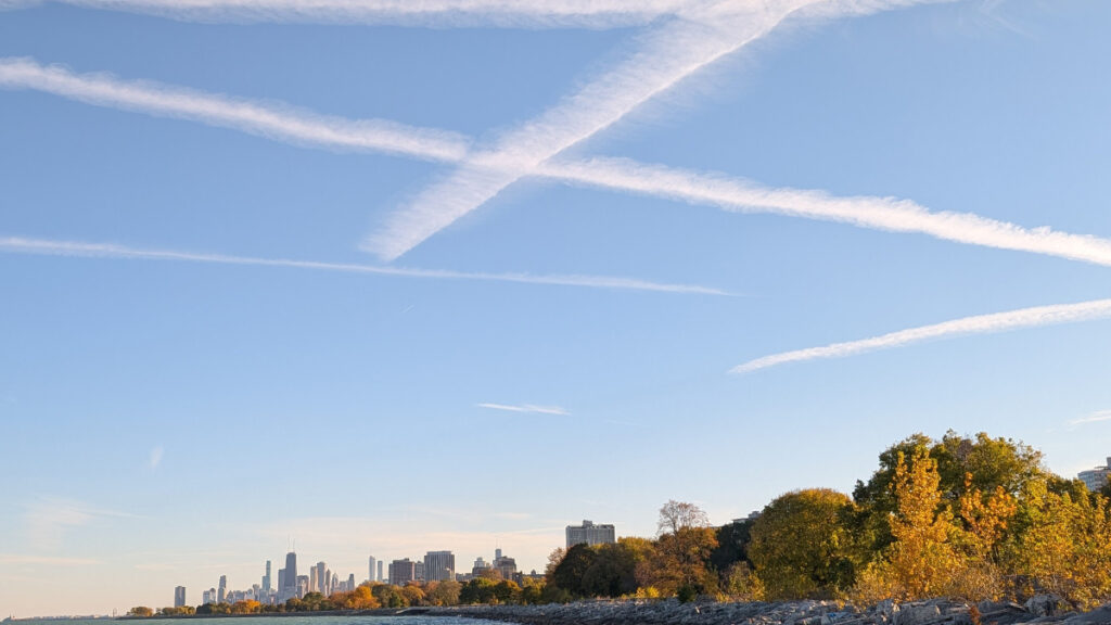 White streaks are seen across a blue sky over a body of water. In the distance is the Chicago city skyline.