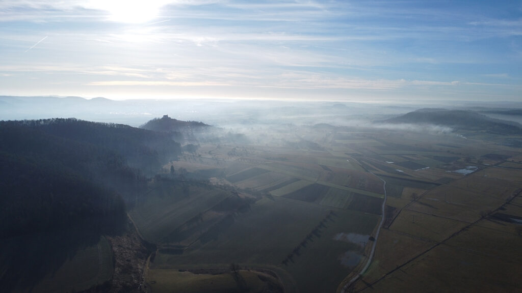 A valley with angular plots of agricultural land is seen from above. The Sun shines through clouds in the background.