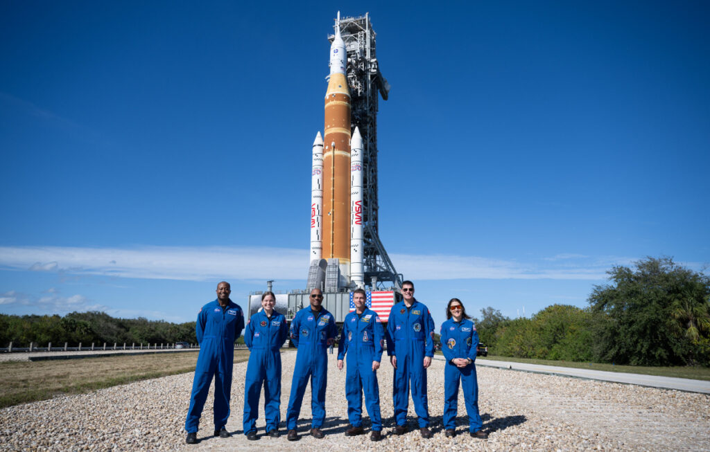 Six astronauts in flight suits stand in front of an orange rocket with boosters on a clear sunny day.