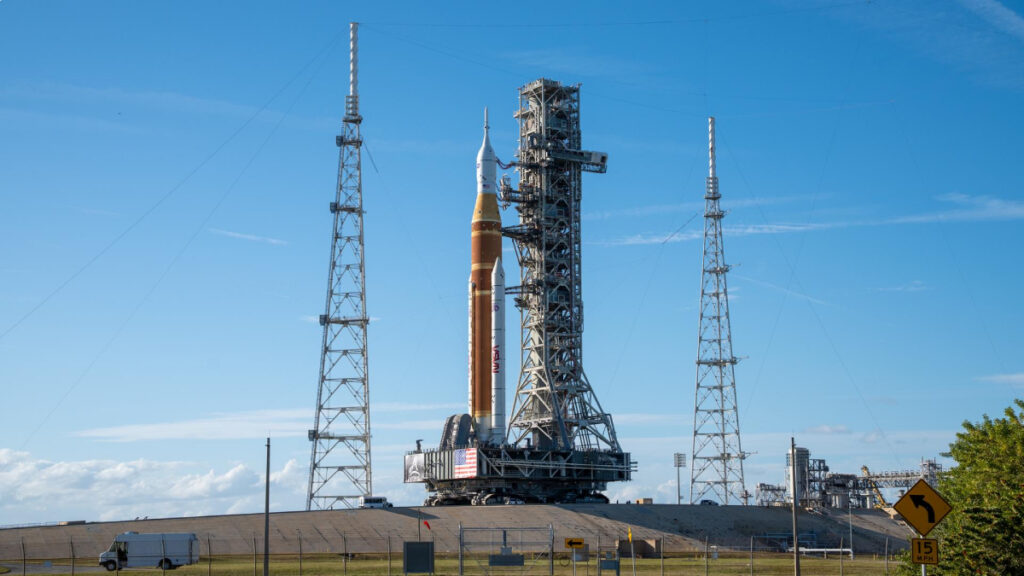 A large platform supporting a rocket and a system of scaffolding is being moved to the launchpad by a crawler-transporter.