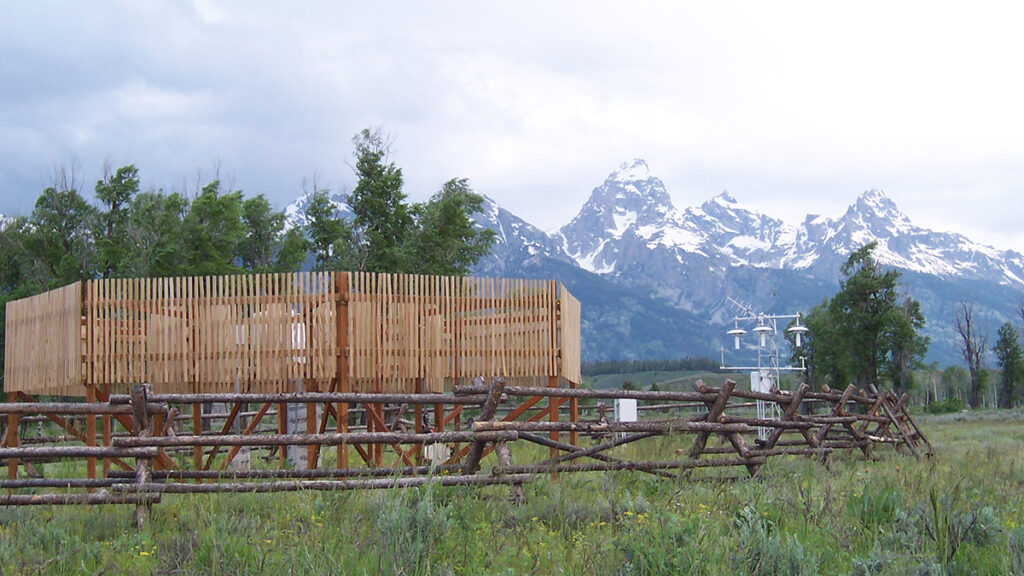 Weather instruments surrounded by a wooden wind shield and rustic lodge pole fencing stand in a grassy clearing with snow-capped mountains in the background.