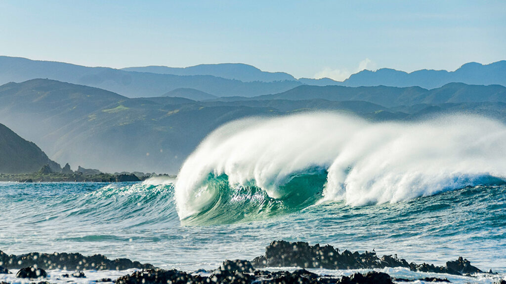 A wave crashes onto a dark, rocky shore. Green rolling hills are in the distance.