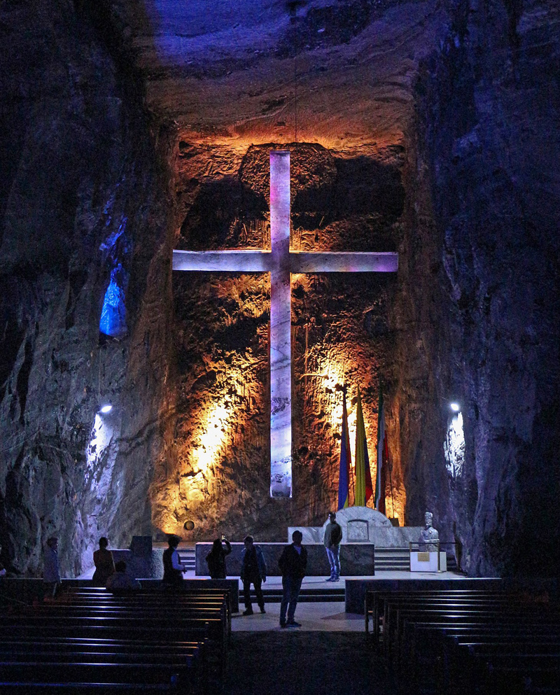 A cathedral built into an underground cavern is dim, but a large cross hanging over the altar is lit with blue and purple lights. A few people stand near the front of the space.