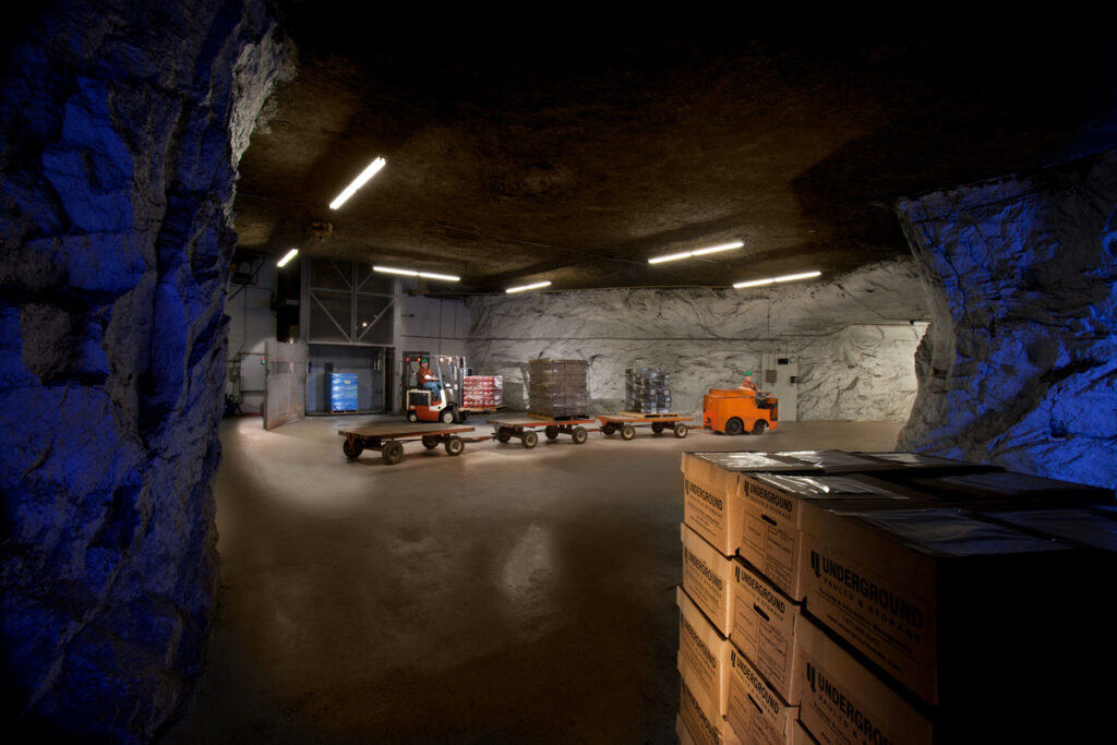 Four cars with wheels are seen in an underground cavern. A stack of boxes is in the foreground, and an elevator leading to the surface is in the background.