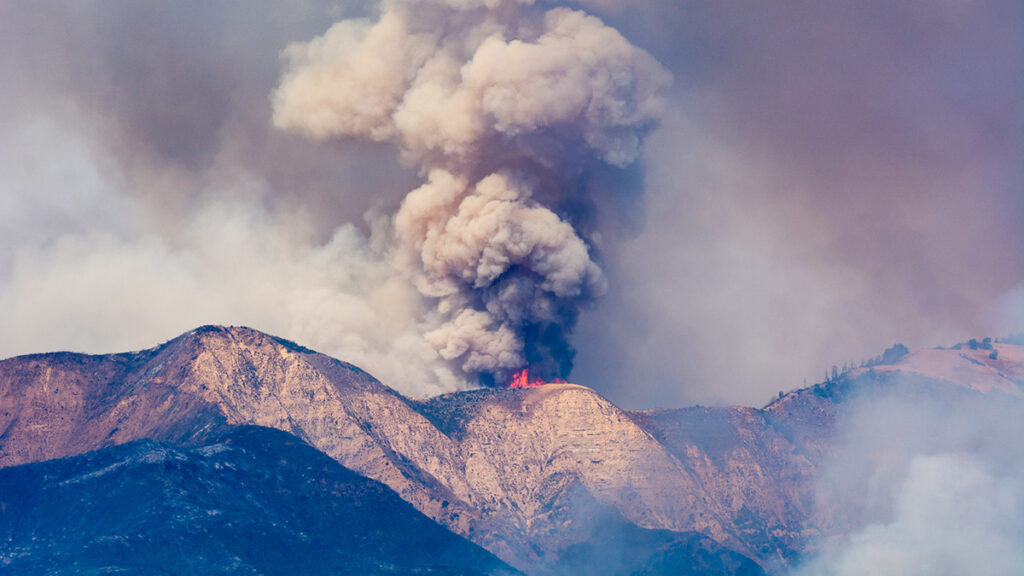 A large gray plume of wildfire smoke rises above a mountain range.