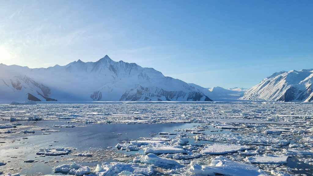 Broken-up ice sits on the surface of an ocean. A snowy mountain, a glacier, and a blue sky are in the distance.