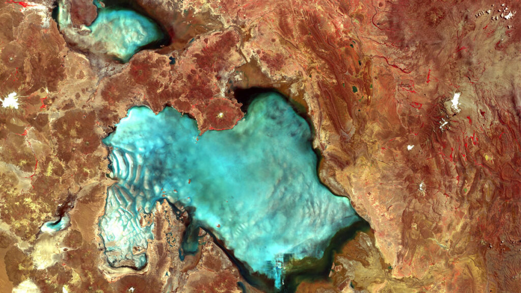 A blue body of water is seen amid a red and brown landscape in an overhead satellite image.