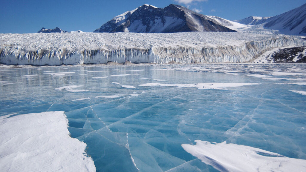 A frozen lake in Antarctica has a blue surface crisscrossed by lines. Behind it is a glacier, a mountain, and a blue sky.