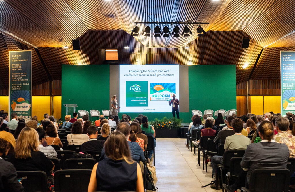Attendees seated in a large hall listen to two presenters standing on a low stage in front of a slide show.